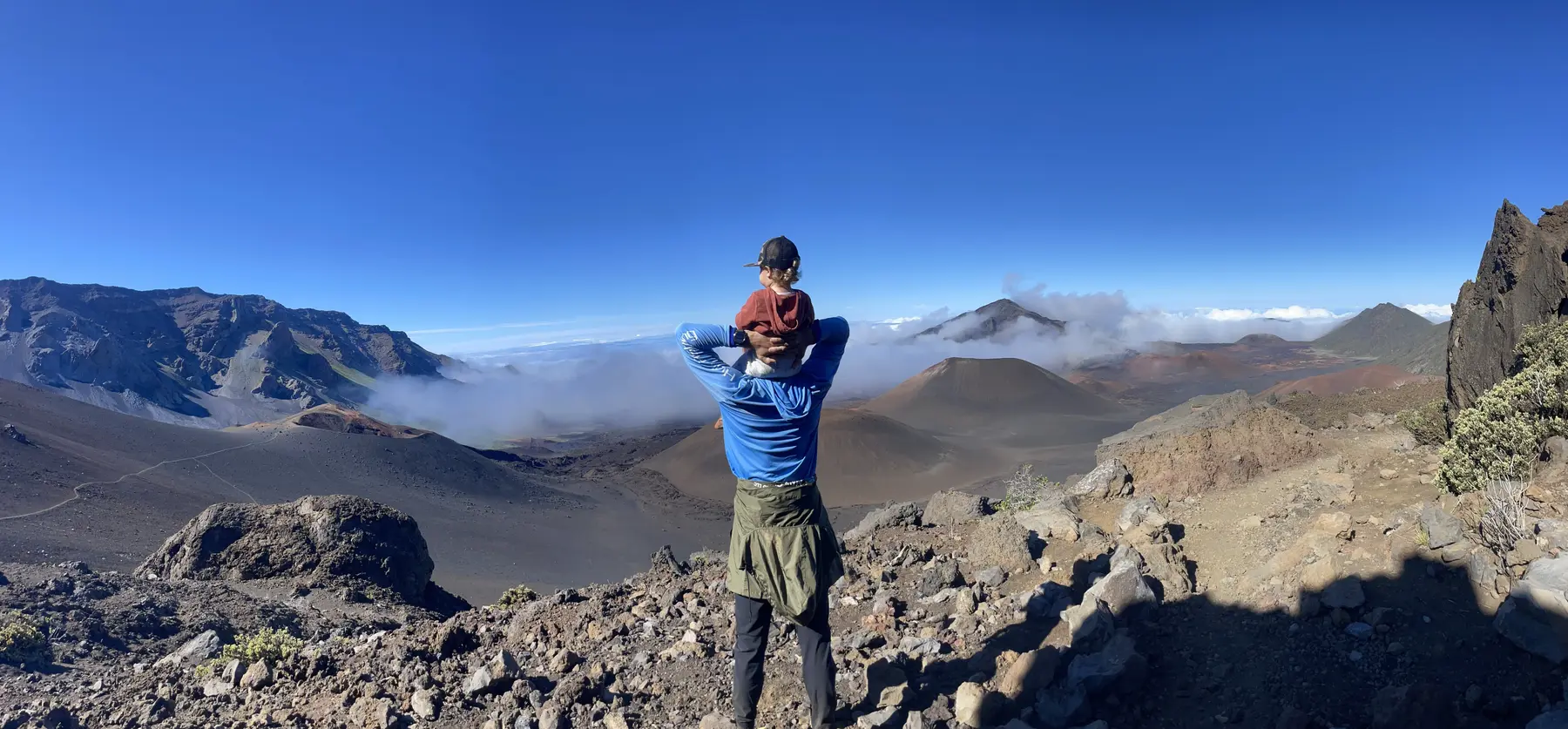 A hiker carrying a small child on their shoulders looking out over the Haleakalā crater on Maui