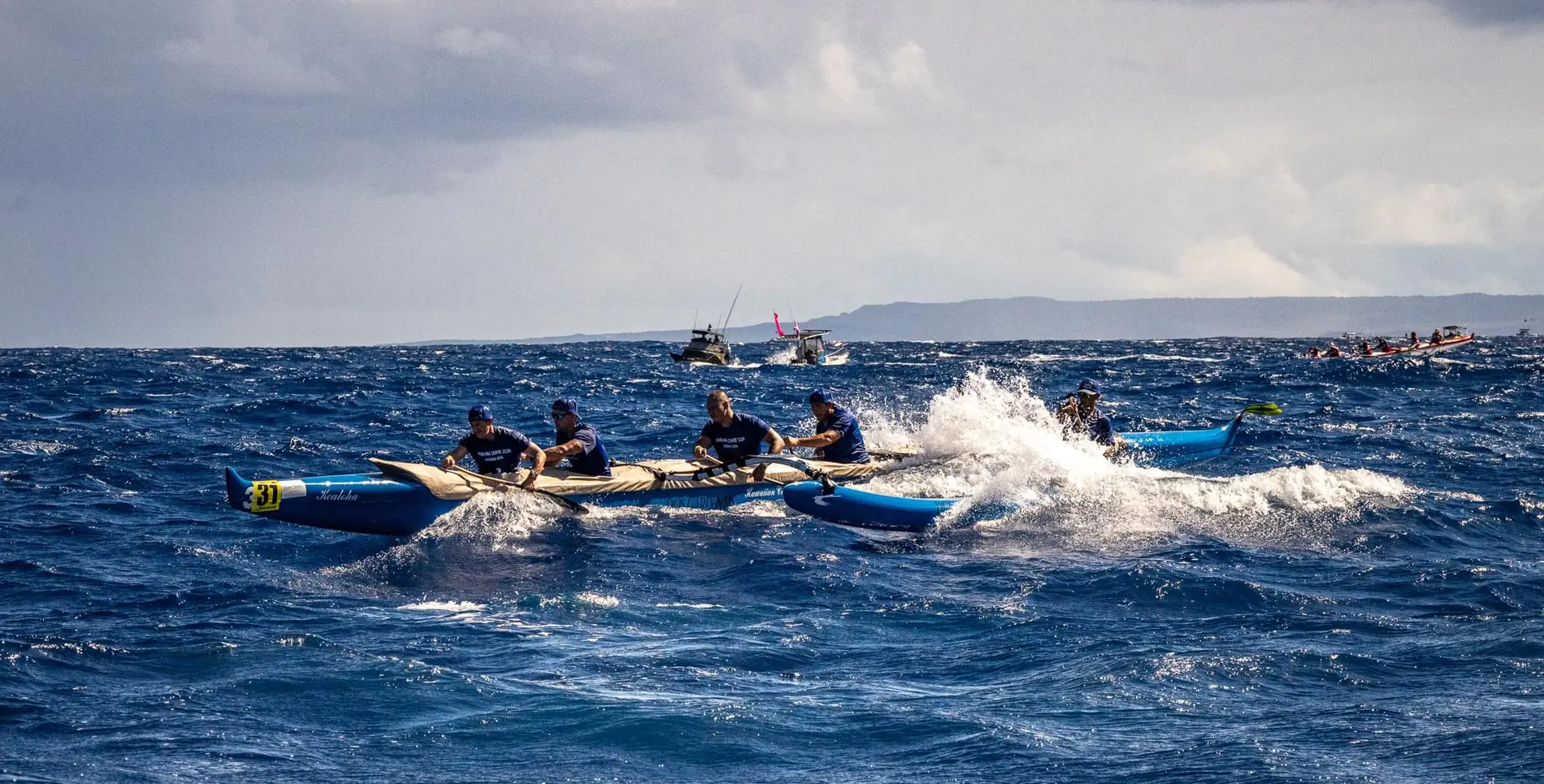Outrigger canoe crew racing through choppy open-ocean swell, with support boats and other canoes behind
